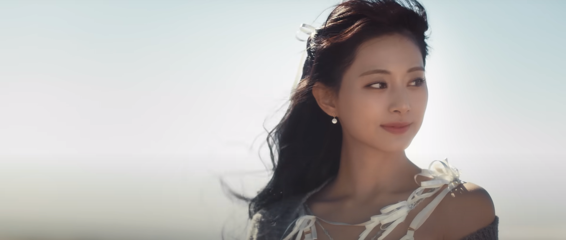 he image features a closeup portrait of a young woman with dark hair styled in a loose, flowing manner, adorned with a white ribbon. she wears a grey offshoulder sweater with white string detailing. her gaze is directed towards the right, and her expression is soft and serene. the background is a blurry, bright sky, suggesting an outdoor setting, possibly a beach. the lighting is natural and soft, highlighting her features and creating a dreamy atmosphere.