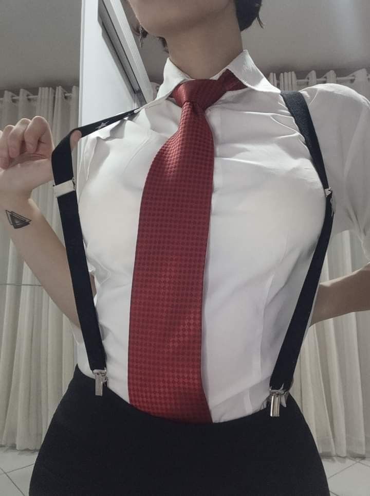a person is wearing a white collared shirt with a red tie and black suspenders. there is a small black triangle tattoo on their arm. the person is also wearing black pants.