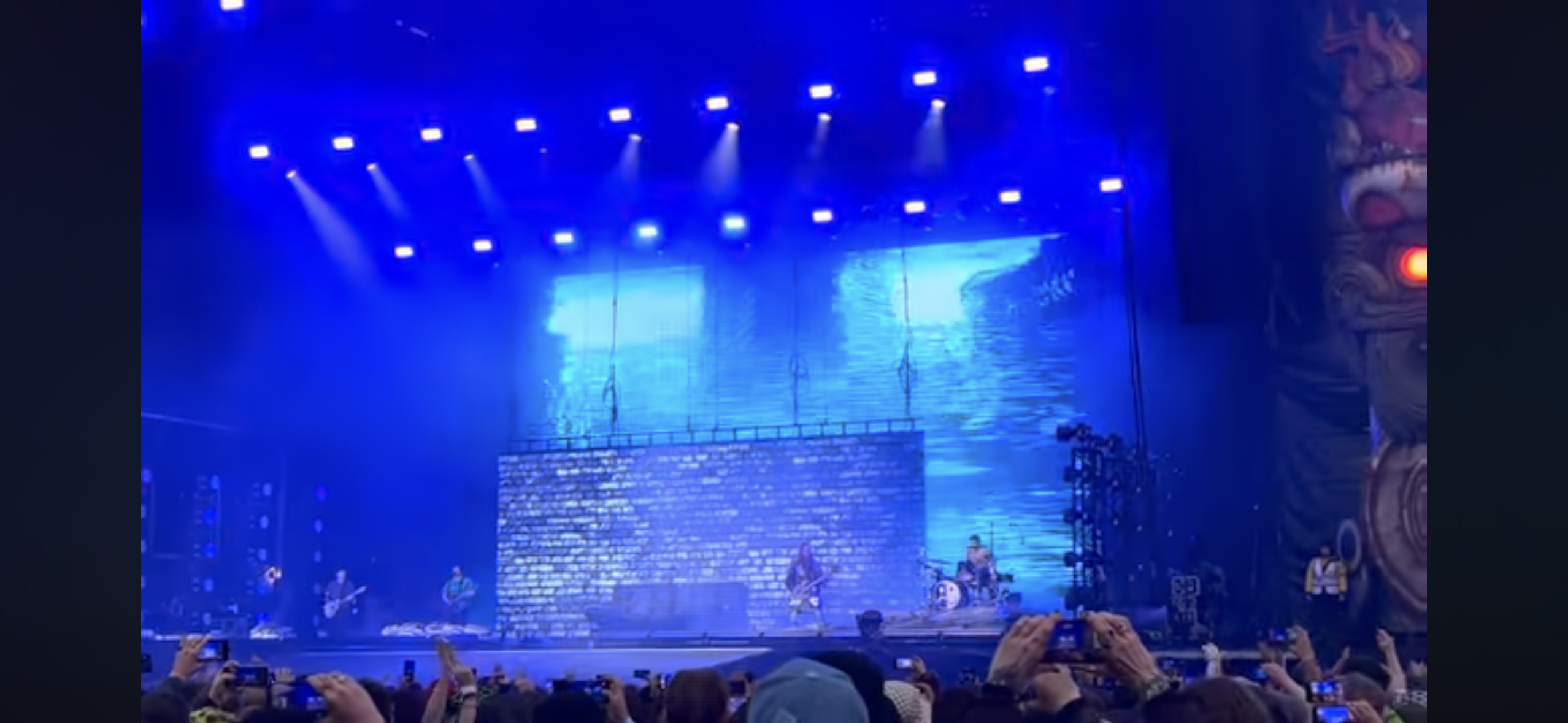 a live concert with a rock band performing on stage under bright blue lights, a crowd of fans in front of them, the band is made up of a drummer, a guitarist and a lead singer.