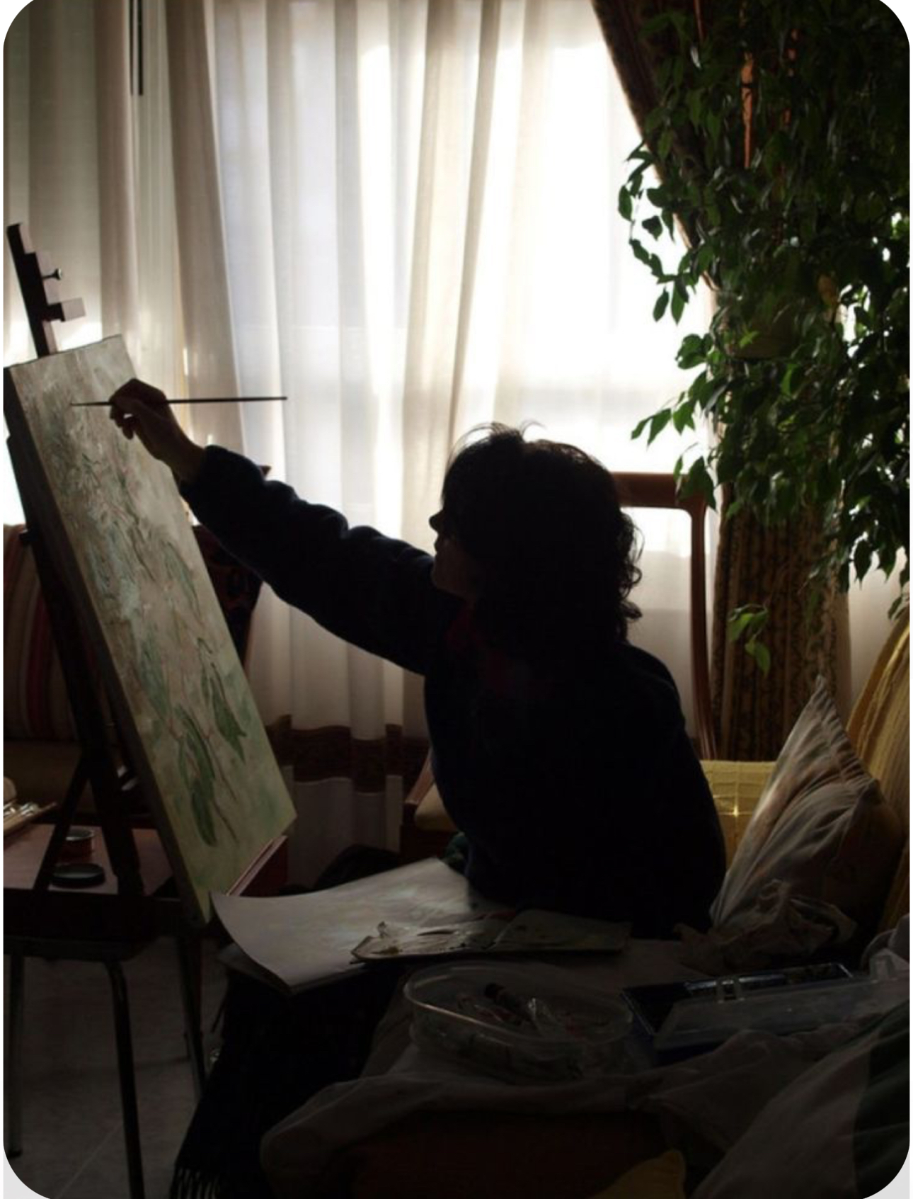 a woman is painting in a naturally lit room. only her silhouette is visible against the light streaming in from the window behind her. she's holding a paintbrush and working on a canvas placed on an easel. beside the easel, there is a table with painting supplies. to the right, a potted plant sits in front of the window. the overall ambiance of the image is calm and creative.