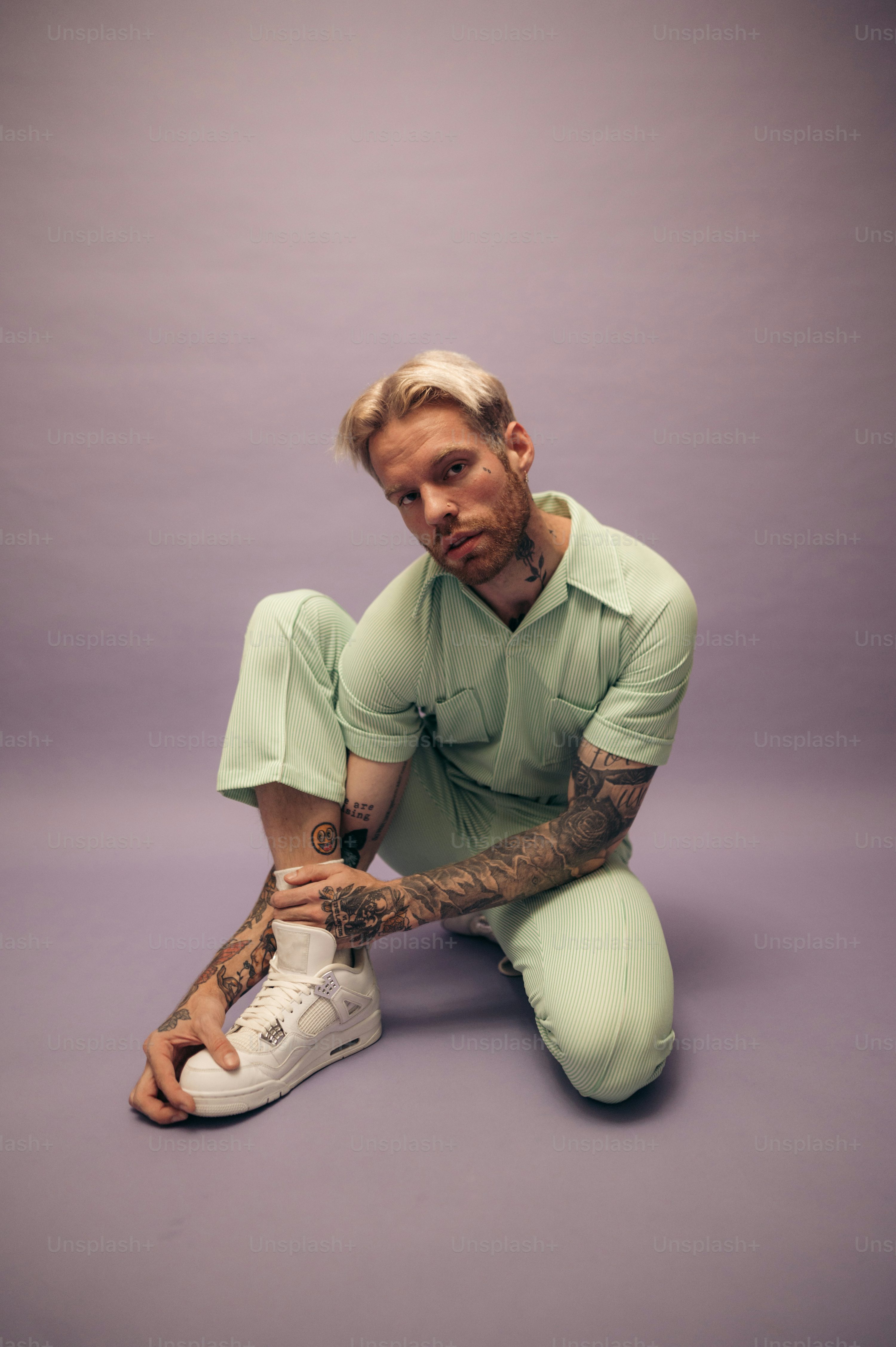 a young man with tattoos sits on a purple studio floor. he's wearing a light green jumpsuit and white air jordan sneakers.  he's looking towards the camera with a somewhat relaxed and cool expression. the overall style is casual and contemporary.