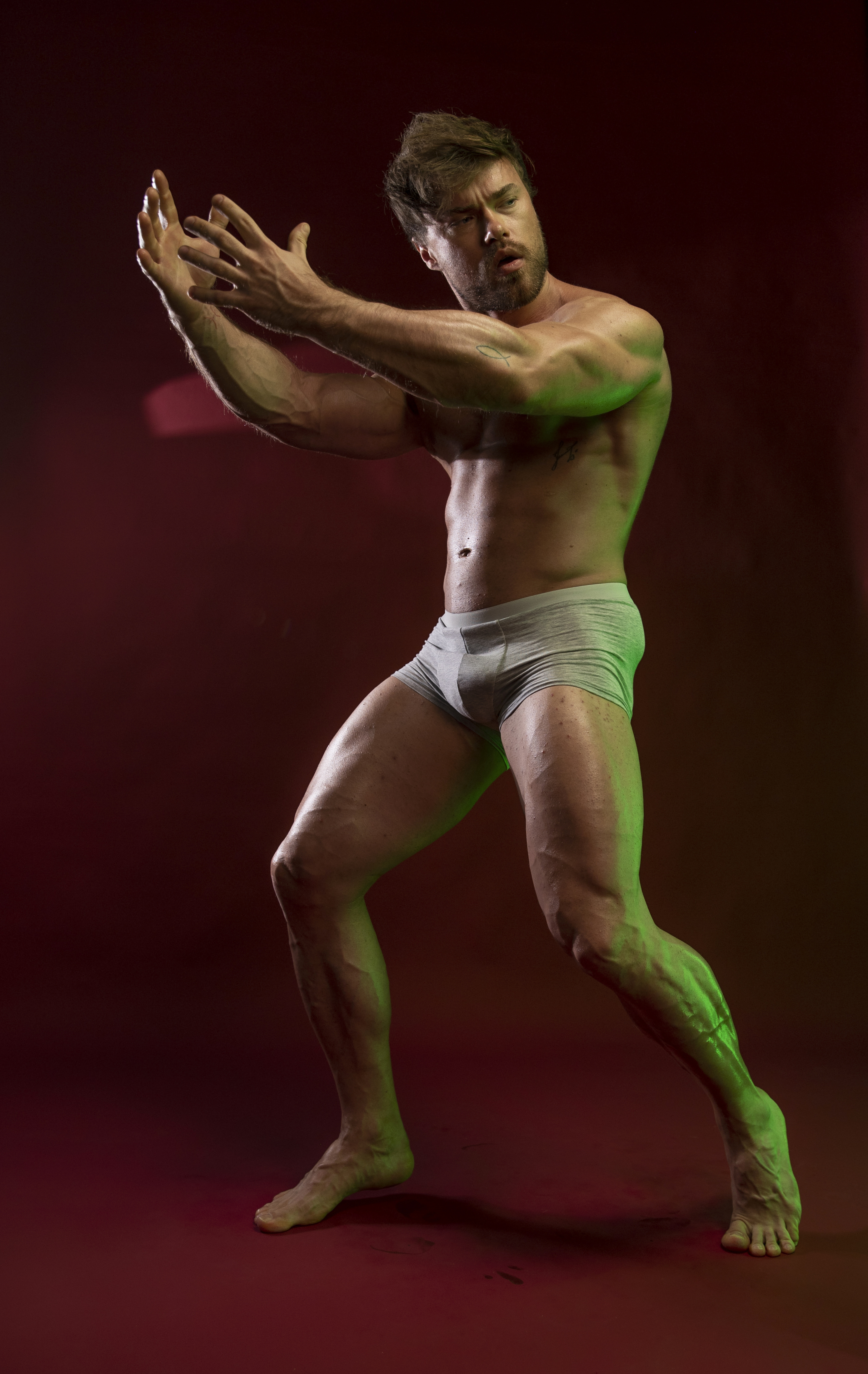 a muscular man in underwear poses barefoot against a red background with green lighting in a studio. he holds his hands up in a gesture.