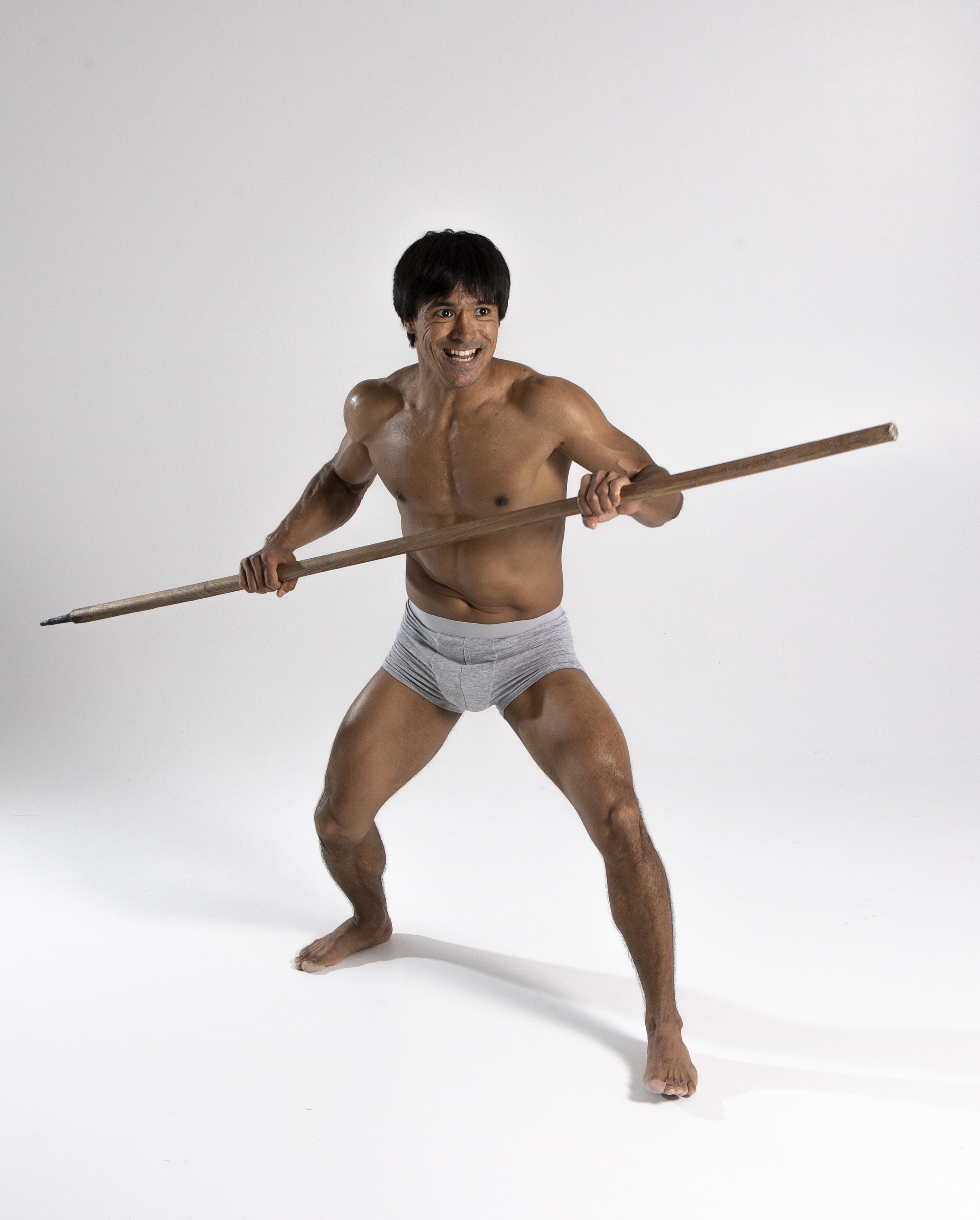 a full shot of a barefoot asian man with short black hair and a muscular build, wearing light gray underwear, posing with a spear in a studio setting. he's shirtless, gripping the spear with both hands, and looks energetic. the background is a plain, light gray.
