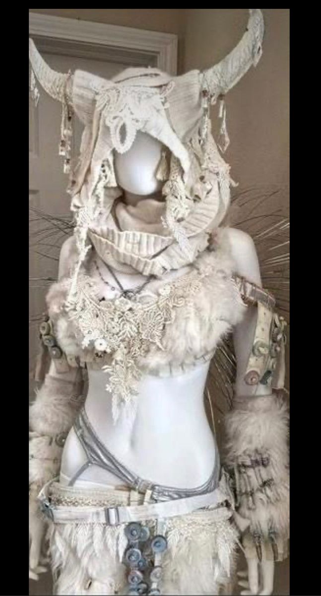 a mannequin is adorned in an elaborate, primarily white costume. the headdress features prominent horns and is decorated with lace and other fabric elements. the costume includes layers of fur, lace detailing around the neck and torso, and a belt with decorative elements. jewelry is visible around the neck, and feathers and other decorative items add to the fantasy aesthetic. the display suggests a fashionforward or artistic presentation of the costume.