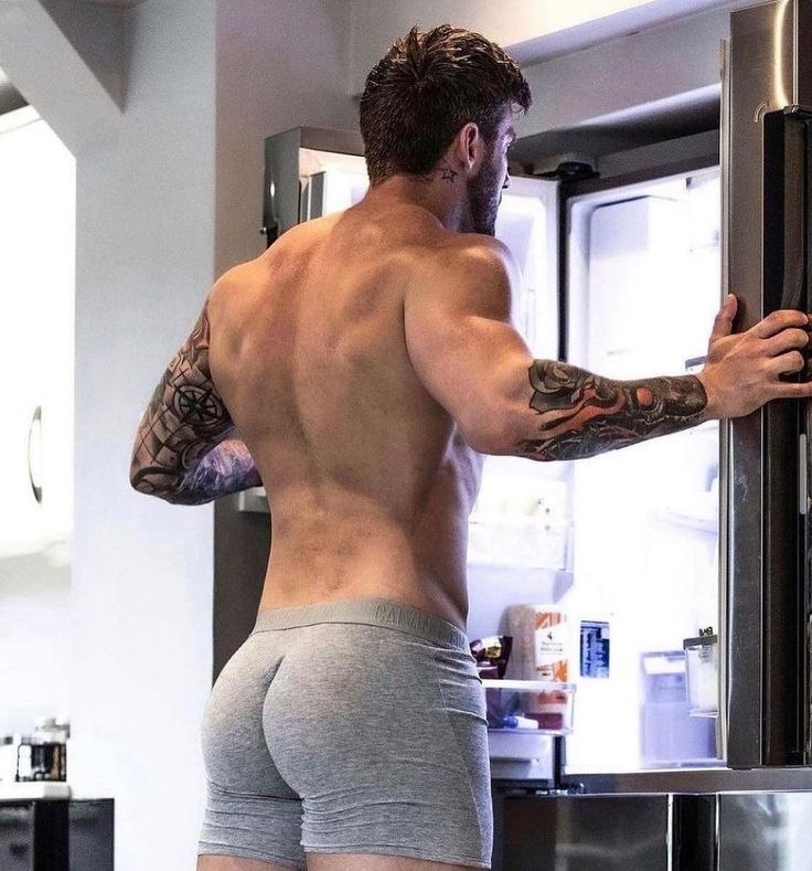 the image shows a muscular, tattooed man from behind, viewed from the waist up. he is wearing grey calvin klein boxer shorts and is standing in a kitchen, opening a refrigerator.  his back is to the camera, showcasing his welldefined physique and tattoos on his arms. the overall mood is suggestive and focuses on the man's body.