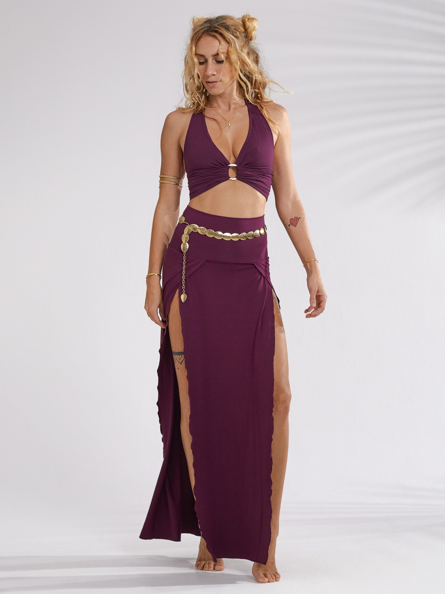 a fullbody shot of a young woman, light skinned with long brown hair styled in a messy bun, is modeling a twopiece outfit. the outfit consists of a dark purple halter top with a gathered front and gold ring detail, and a matching floorlength maxi skirt featuring high slits on both sides. she's also wearing a gold coin belt and has minimal jewelry. the woman's pose is relaxed, and the background is a simple, light gray, providing a clean backdrop to highlight the attire. the overall style is bohochic or festivalready.
