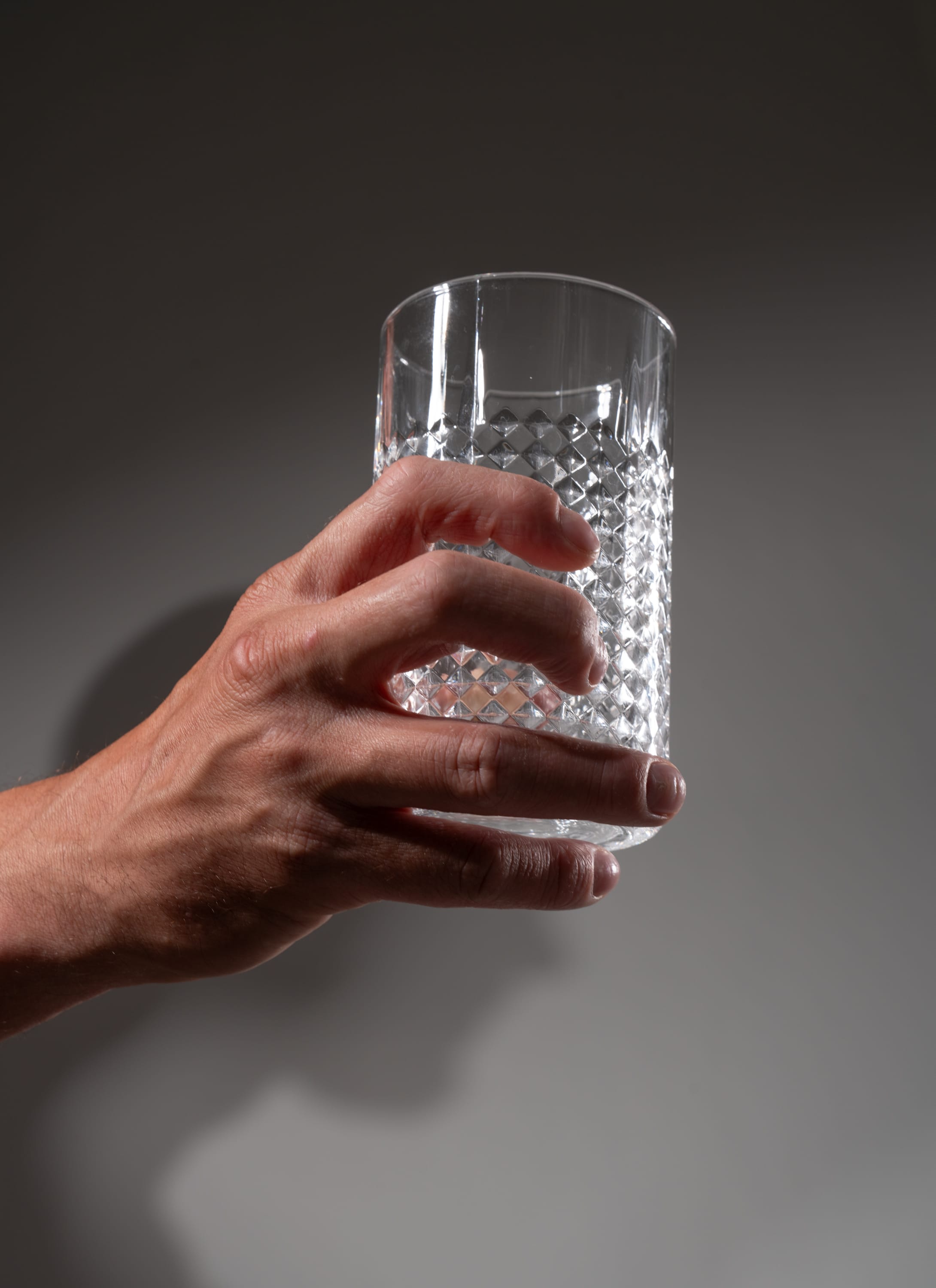 a hand holds a clear, empty glass with a textured design against a gray background. the glass is illuminated by studio lighting, highlighting its transparency and texture.