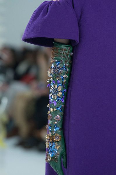 loseup view of a woman's arm wearing a long, darkgreen glove heavily embellished with various colored beads and jewels. the glove is intricately decorated with florallike designs and shimmering elements.  the woman is wearing a rich purple dress with a puffed sleeve. the background is blurred, suggesting a fashion show or photoshoot setting. the focus is sharply on the glove, showcasing the craftsmanship and detail of the embellishments.