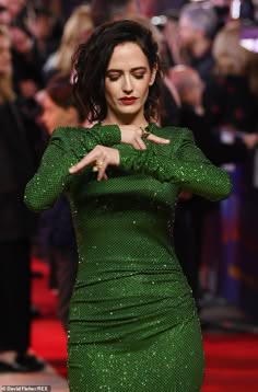 a woman wearing a green sequined dress is posing on a red carpet. she has her hands up near her face and is smiling. there are other people in the background. this image was taken at an awards ceremony or other event where celebrities are often seen.