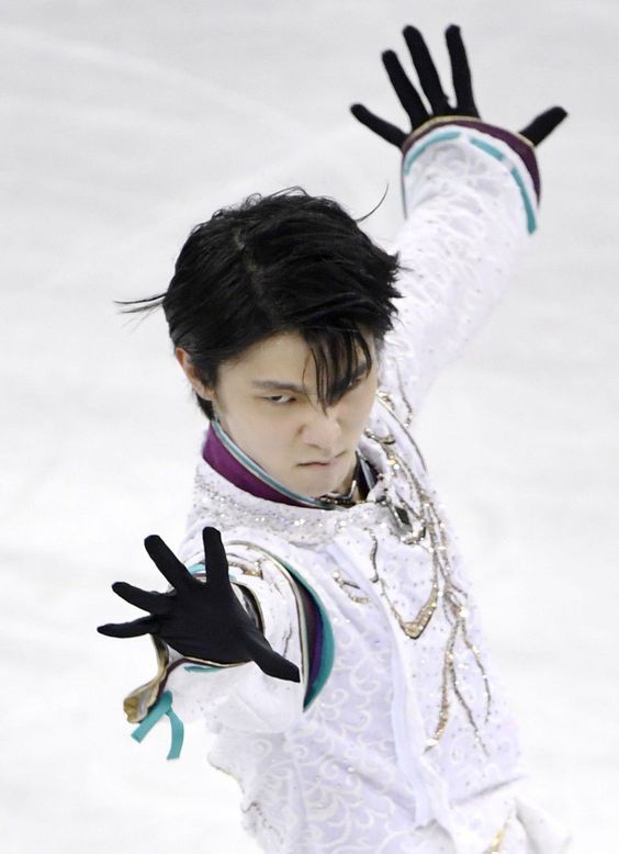 a figure skater in midair, his arms outstretched and his face contorted in concentration. he is wearing a white and blue costume with black gloves. the ice is visible in the background.