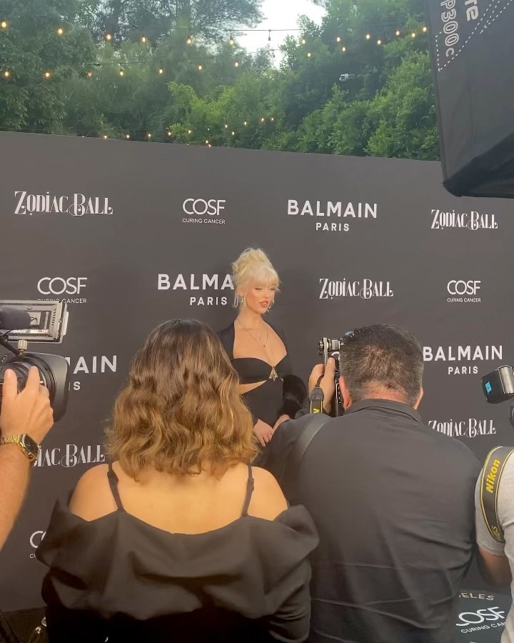 a woman with blonde hair is being photographed on a red carpet at an event. she is wearing a black dress and standing in front of a backdrop with the logos for "zodiac ball," "cosf curing cancer," and "balmain paris." several photographers are positioned around her taking pictures with professional cameras.