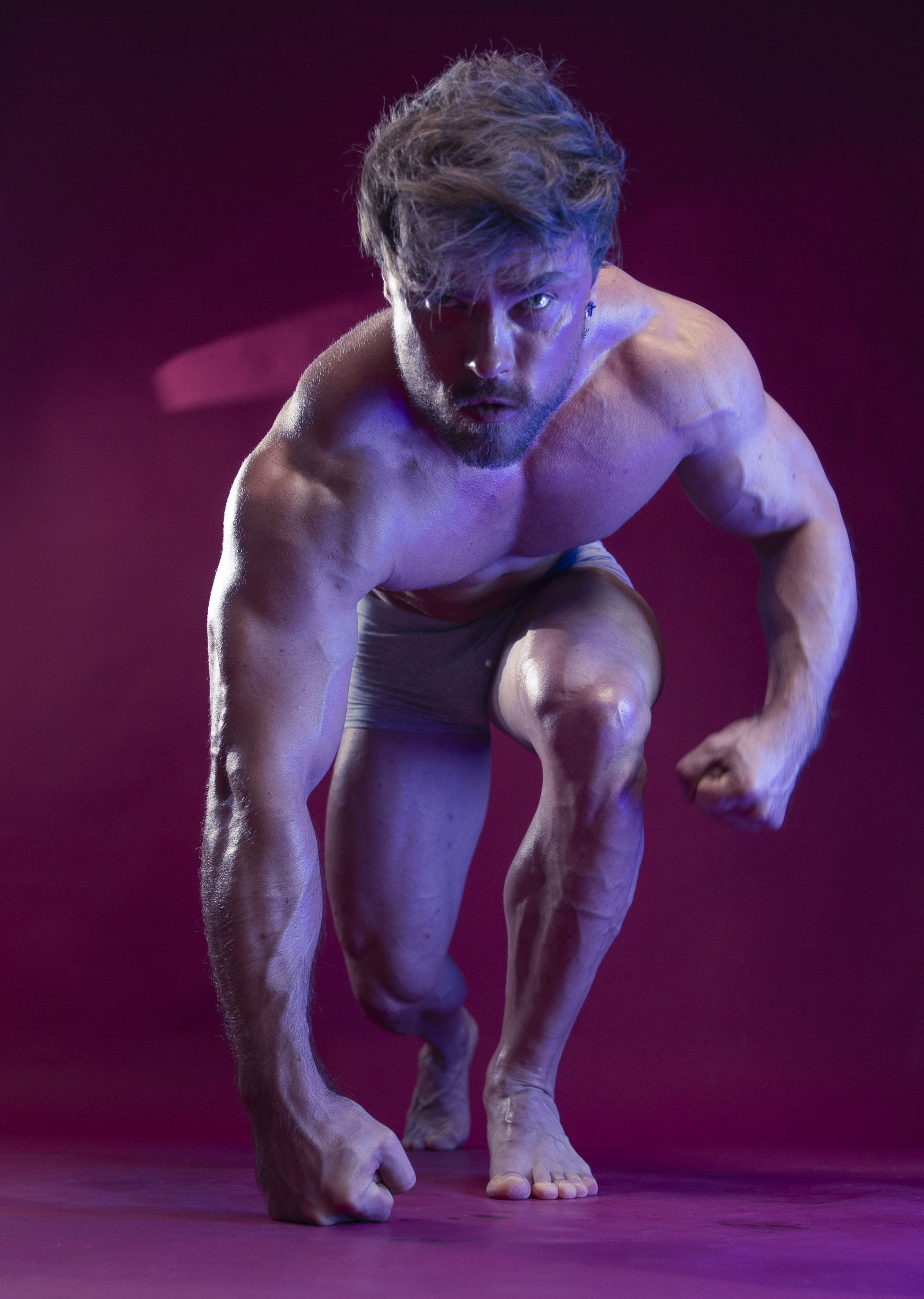 the image shows a muscular man in a crouching position, as if preparing to spring forward. he is barechested and wearing only briefs. his physique is welldefined, showing prominent muscles and visible veins. the lighting is dramatic, with purple and pink hues highlighting his form. the man's expression is intense and determined, suggesting strength and focus. he is positioned indoors against a solid background, likely in a studio setting.
