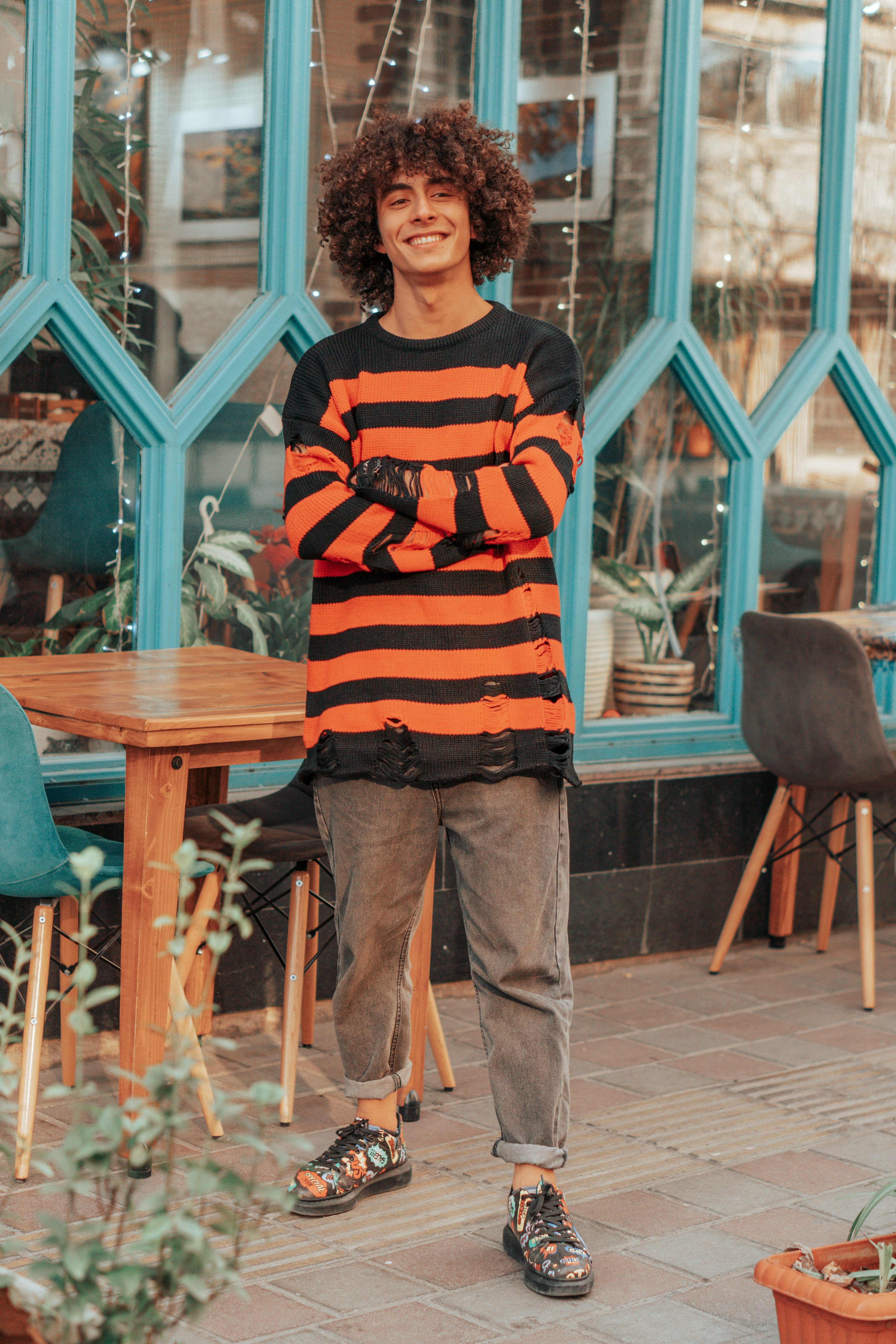 a full shot shows a young man standing outside of a cafe with his arms crossed. he has curly brown hair and a warm smile. he wears a black and orange striped sweater with distressed details, paired with grey jeans cuffed at the ankles, and patterned shoes. behind him is a cafe with blue window frames. a wooden table and chairs are placed beside him.