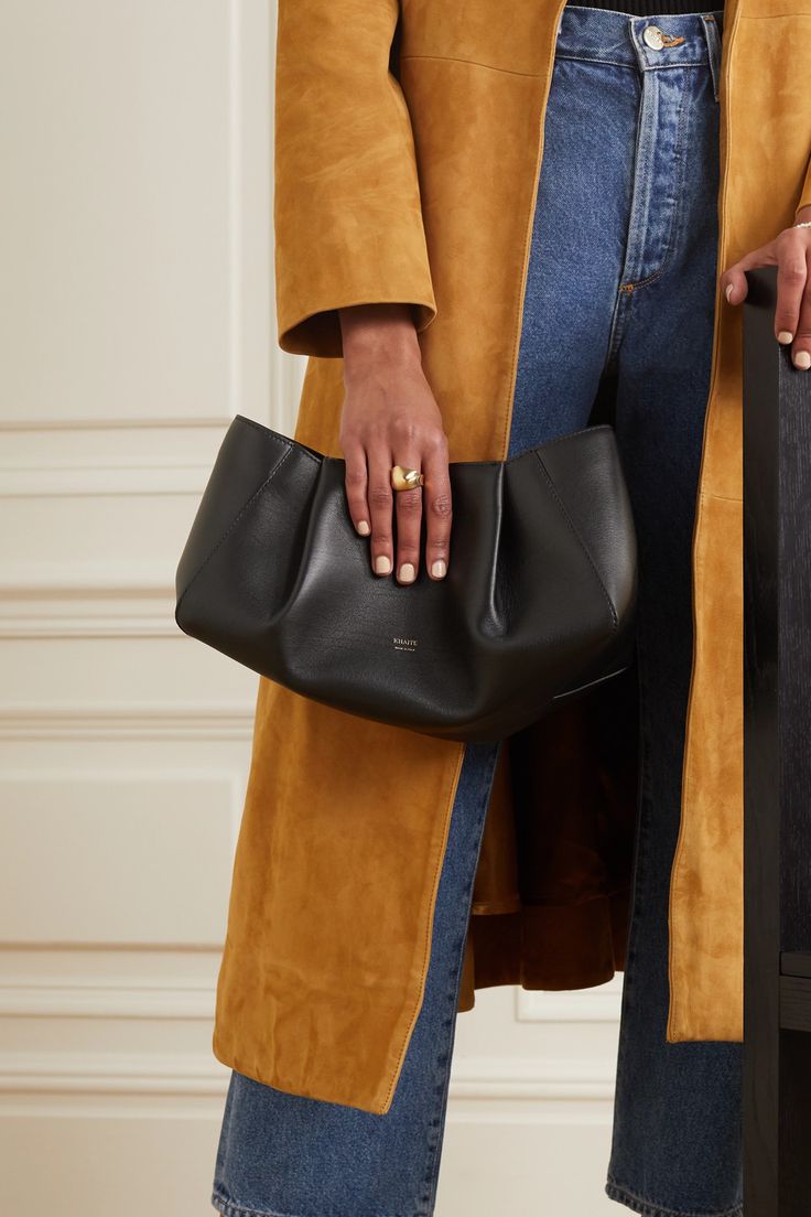 a woman is shown holding a black leather pleated clutch purse. she is wearing a mustard yellow suede coat and dark wash denim jeans. she is also wearing a gold ring on her left ring finger. the image is welllit and the focus is on the woman and the handbag.