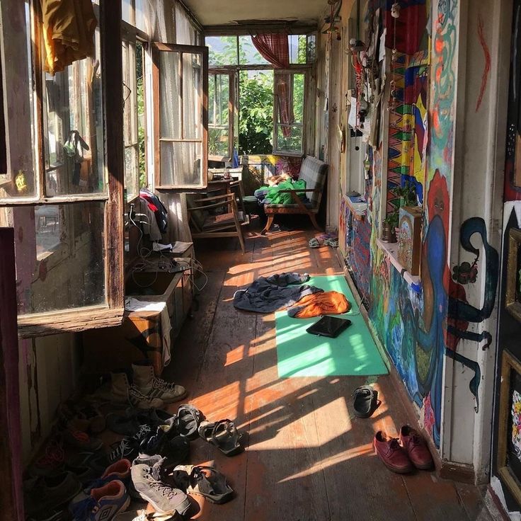 the image depicts a sunlit porch or veranda with a distinctly bohemian and eclectic style. the wooden floor is worn and shows its age, and the walls are covered in vibrant, colorful graffiti and artwork. numerous items are scattered about, including a pile of shoes, clothes, and a laptop on a green yoga mat. an old armchair is visible, along with overgrown plants visible through the aged windows.  the overall atmosphere is relaxed and cozy, characterized by a slightly cluttered yet artistic aesthetic. sunlight streams in through the windows, illuminating dust motes and creating interesting shadows on the floor.  the space feels livedin and personal, reflecting the personality of its inhabitant.