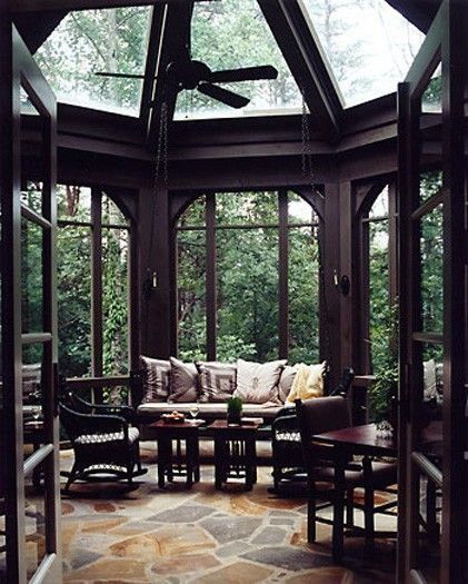 this is a photo of a sunroom. the sunroom has dark wood framing and glass windows. there is a seating area with a sofa and cushions, a table, and chairs. the room also has a ceiling fan. the floor is made of stone. the windows look out onto a forest.