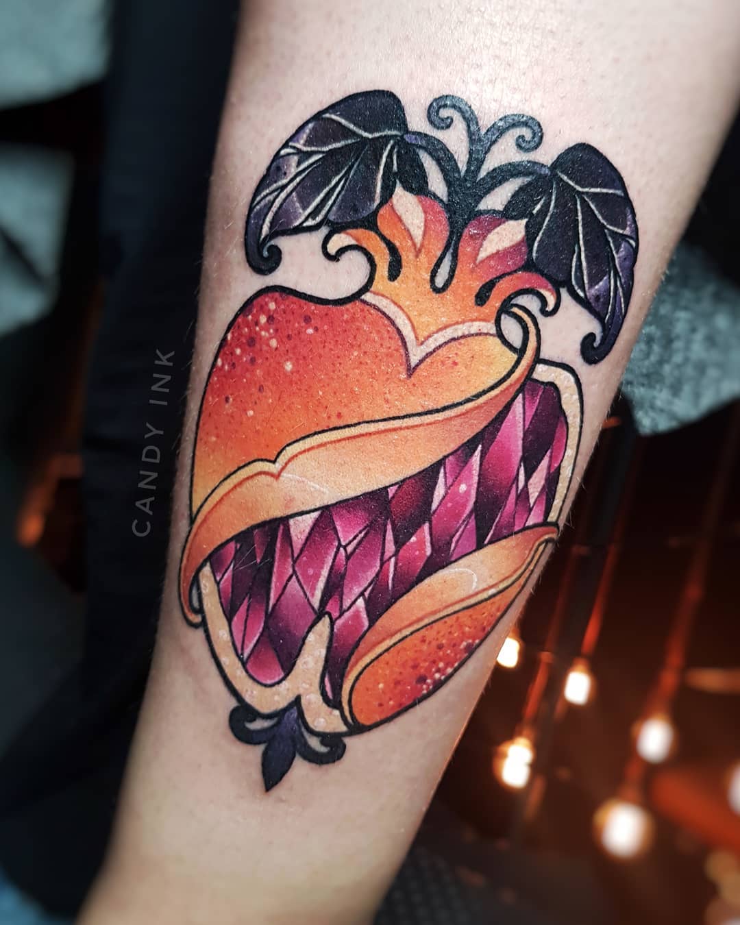 a tattoo on a person's arm. the tattoo is of a stylized pomegranate with a heart shape. the outer layer of the pomegranate is orange and yellow, while the inside is made up of pink and purple crystals. there are black leaves with white highlights at the top of the pomegranate. the tattoo is done in a neotraditional style with bold lines and shading.