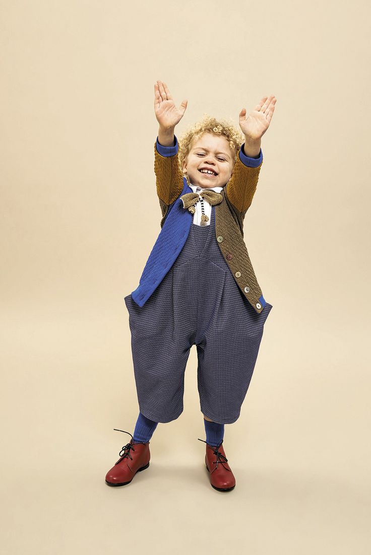 a full body shot of a young boy with curly blonde hair smiling and holding his hands up in the air. he is wearing blue overalls, a brown and blue jacket, a bowtie, and red shoes. he is standing in front of a plain beige background.