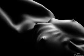 the image is a black and white closeup photograph of a nude female torso. the lighting is dramatic, using strong chiaroscuro to emphasize the curves and textures of the body.  the model is lying down, and the focus is primarily on the upper body, highlighting the subtle folds and shadows of the skin.  the overall mood is sensual and intimate, with an artistic and abstract feel. the image emphasizes form and texture more than precise detail.