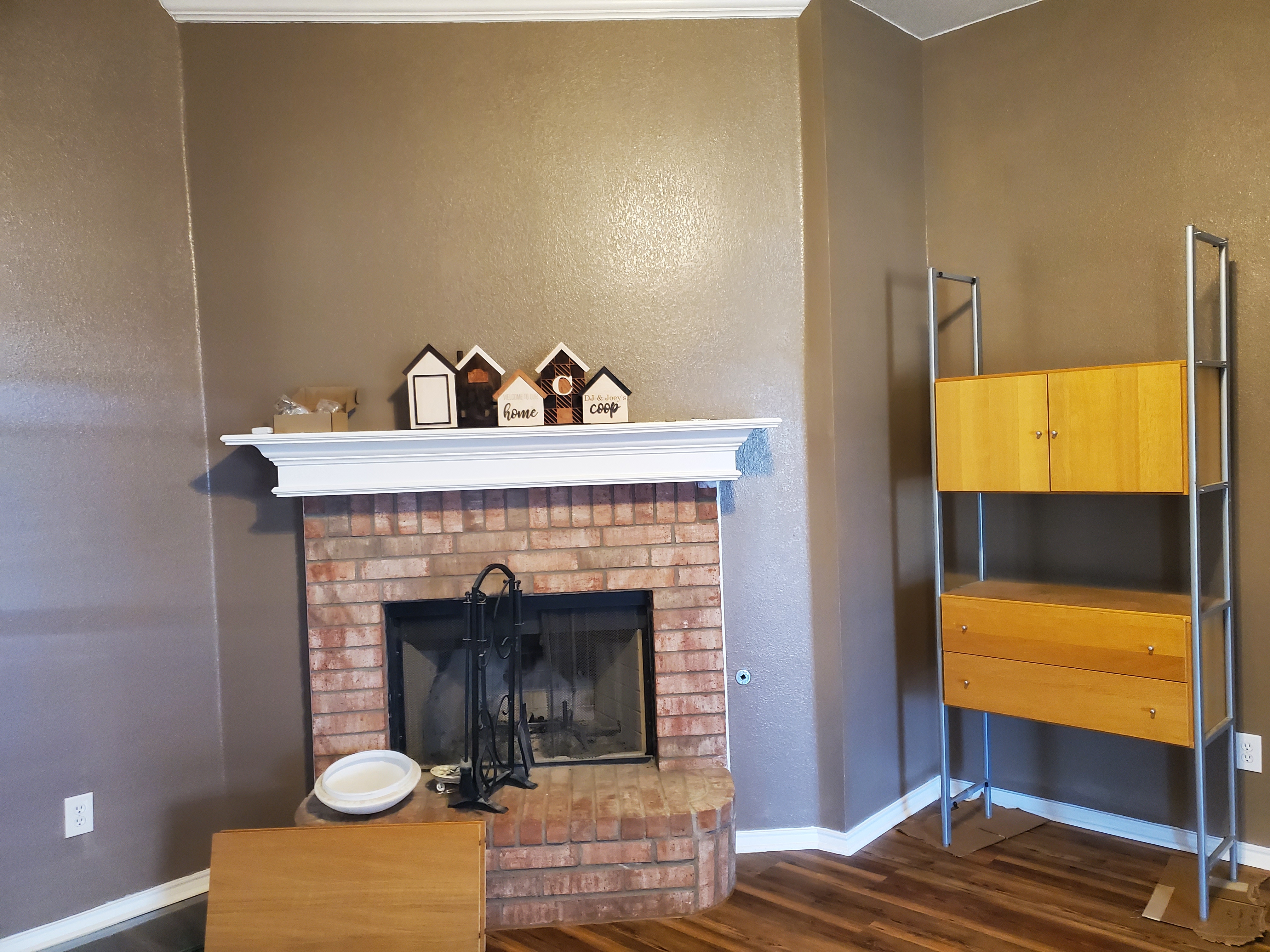 the photo shows an interior shot of a living room featuring a brick fireplace with a white mantel, on which are decorative wooden houses. a shelving unit with lightcolored wood shelves and silver frame stands to the right of the fireplace. there are wood floors, and the walls are brown. a table sits in front of the fireplace.