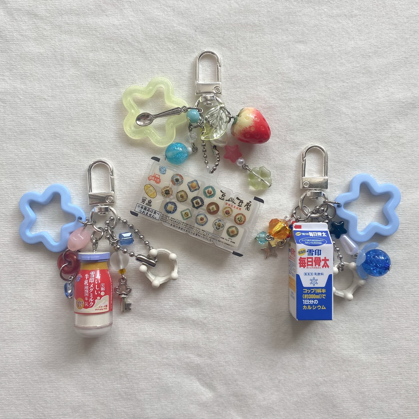 the image shows three handmade keychains lying on a white surface. each keychain features a mix of charms including miniature food items, medicine bottles, miniature snack packets, and milk cartons, all attached to a metal keychain clip. each keychain also includes a starshaped decorative plastic frame of different colors and various decorative beads.