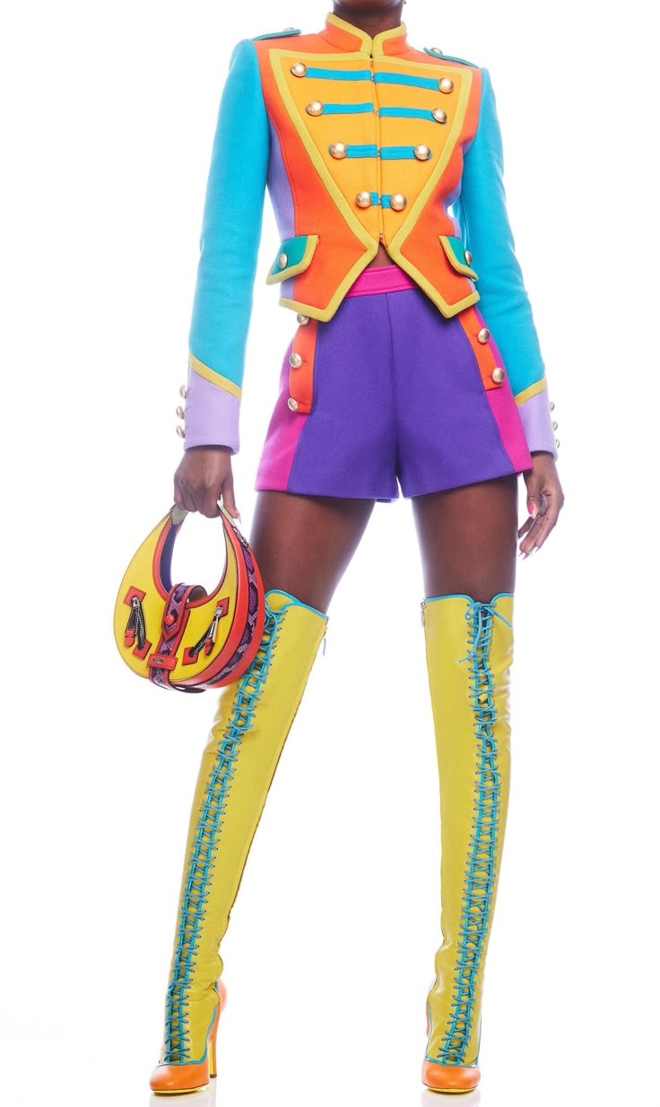 a woman stands against a white background wearing a colorful outfit including a cropped jacket, shorts, kneehigh boots, and carrying a handbag. the jacket is a mix of turquoise, orange, and gold with rows of buttons, resembling a militarystyle jacket. the shorts are colorblocked with purple and pink. the boots are yellow with light blue lacing. the handbag is yellow with red and black details.