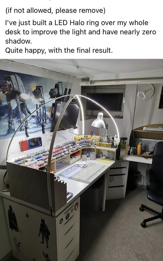 the image shows a wellorganized desk with a custombuilt led halo ring for improved lighting and minimal shadows. the desk has various tools, paint bottles, and a monitor. a desk lamp provides additional light. the background features wall art with atat walkers. a clock is visible on the wall. the setup includes an office chair and drawers for storage. the desk also has an organizer for paint bottles and other supplies. a window is visible in the background. the desk setup appears to be a hobby space.