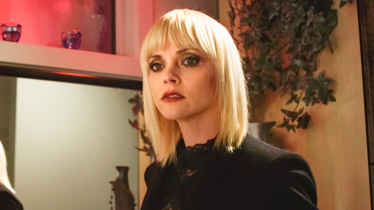 loseup portrait of a woman with a blonde bob haircut and bangs. she is wearing a black jacket over a black lace top. the setting appears to be indoors, dimly lit with red lighting in the background. the woman has a serious and somewhat mysterious expression on her face. the overall style is elegant and fashionable, suggestive of a still from a television show or film.