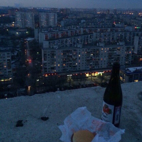 the image shows a rooftop view of a dimly lit city at night. in the foreground, there is an open paper bag containing a burger and a bottle of wine. the majority of the image is filled with a vast expanse of residential buildings, with their windows illuminated against the dark sky. the perspective suggests a sense of solitude and quiet observation of the city below.