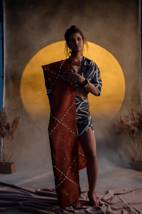 a full shot of a woman standing against a backdrop of a large, yellow circle. she is wearing a stylish outfit that appears to blend traditional and modern elements. the clothing consists of a dark top with white geometric patterns, a short skirt in a similar pattern, and a long terracottacolored shawl or wrap that drapes elegantly around her. her jewelry includes a necklace and earrings. the overall mood is one of elegance, confidence, and artistic flair. the lighting and the background create a dramatic yet intimate atmosphere.