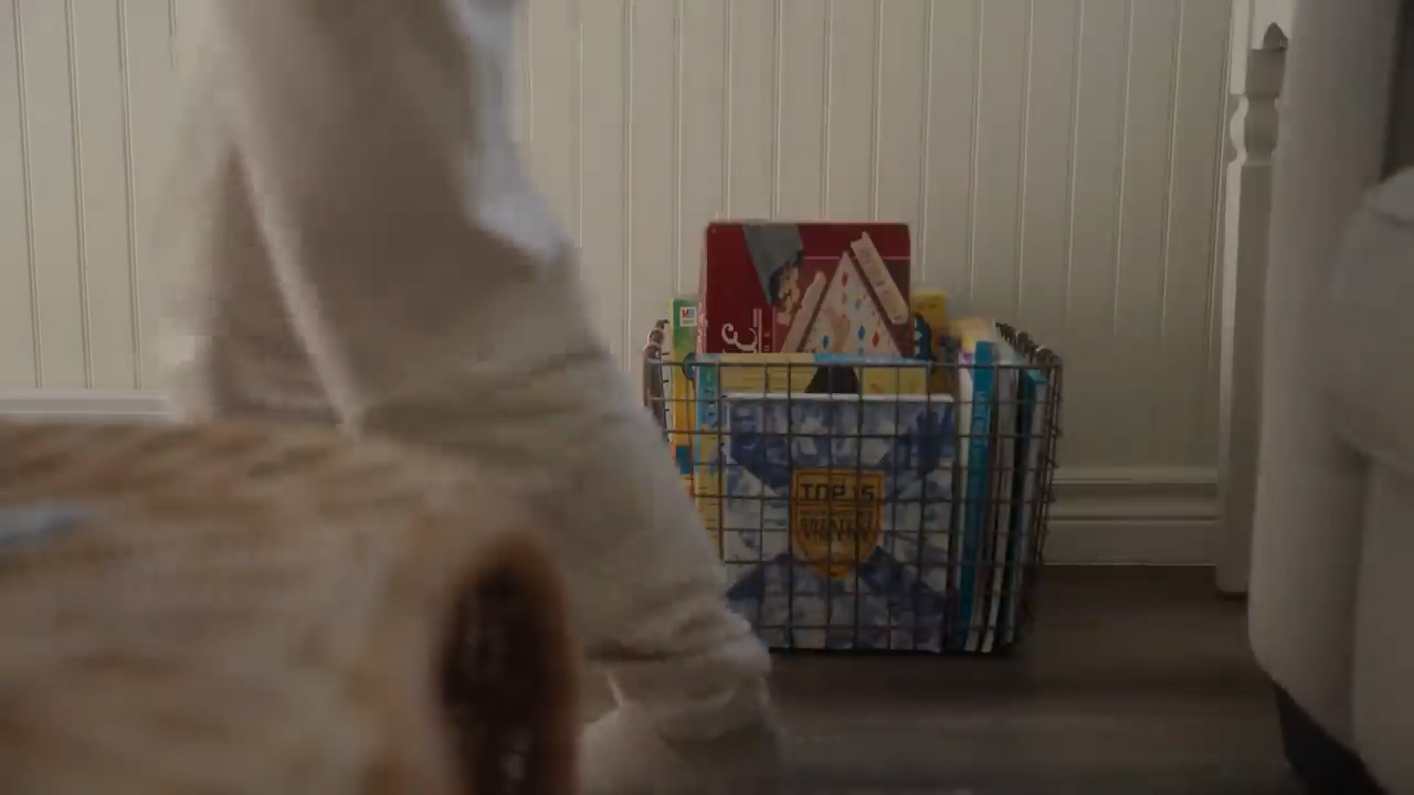 a young child walks past a basket of books in a bright, naturally lit bedroom. the child is wearing white pants and is out of focus, while the basket of books is in focus in the foreground. the bedroom has white walls and a lightcolored wood floor.