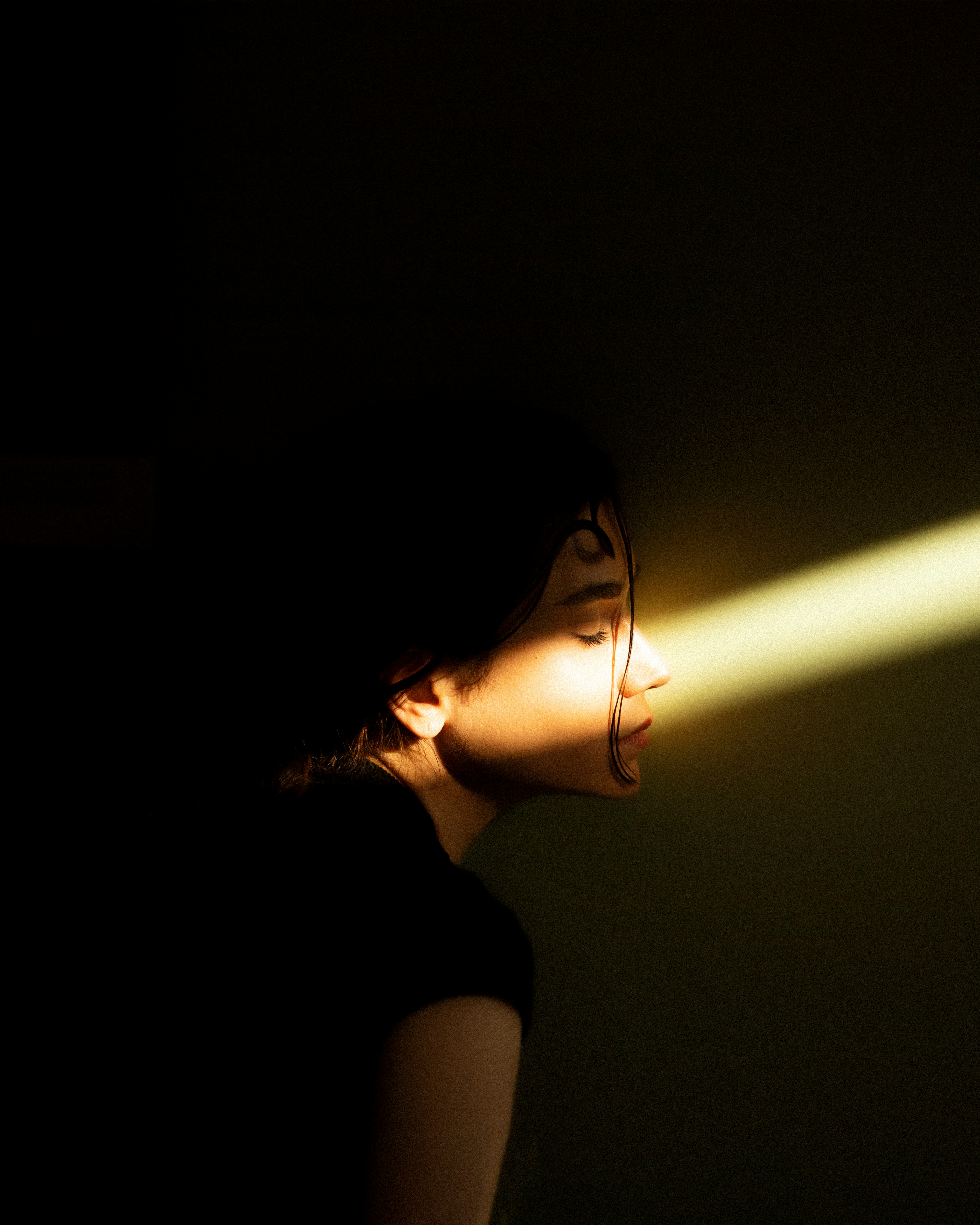 a woman is partially illuminated by a bright beam of light against a dark background. her eyes are closed, and half of her face is bathed in the warm light. the rest of the image fades into darkness, creating a strong contrast.