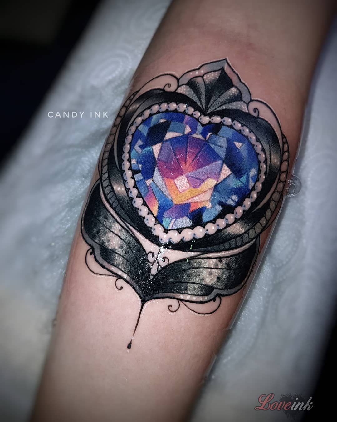 a tattoo of a heartshaped jewel on someone's inner forearm. the jewel is blue, purple, and pink, and is surrounded by a border of white dots and black and gray filigree. there are also sparkles around the jewel. the tattoo is by candy ink and love ink.