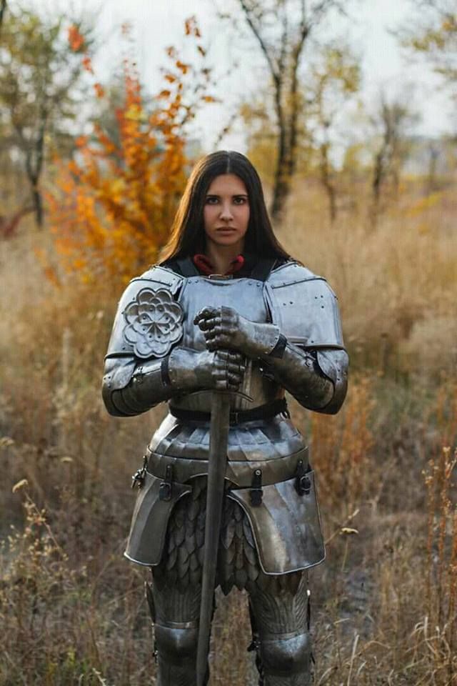 a woman in full plate armor stands in a field of tall grass. she is holding a sword in her hands and looks determined. the armor is silver and the woman's hair is long and dark. there are trees in the background.  the woman is dressed as a knight or warrior from the middle ages.