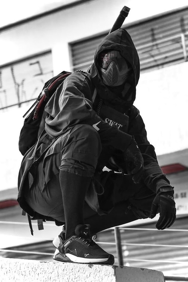 a person in a tactical gear with a mask on is crouching in an urban environment. the image is in black and white and the person is wearing a black and white clothing with a black backpack. they are crouched behind a metal railing. the person is looking into the camera and it looks like they are ready to take action.