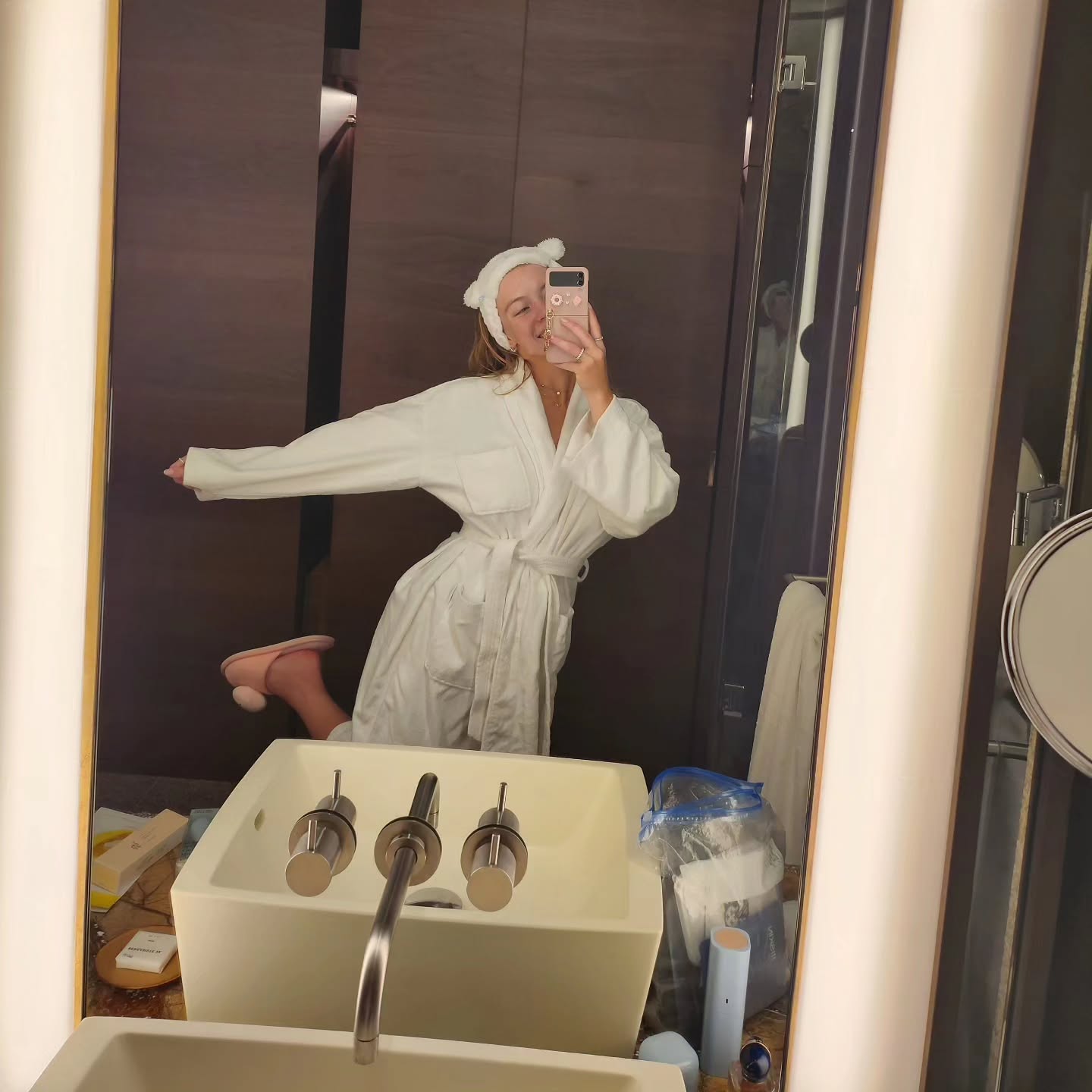 a young woman in a white bathrobe is taking a selfie in a bathroom mirror. she's wearing a cute bunny ears headband and pink slippers.  the bathroom is modern and luxurious, featuring a sleek sink and faucet.  various toiletries and items are visible in the background, suggesting a hotel or spa setting. the overall mood is one of relaxation and selfcare.