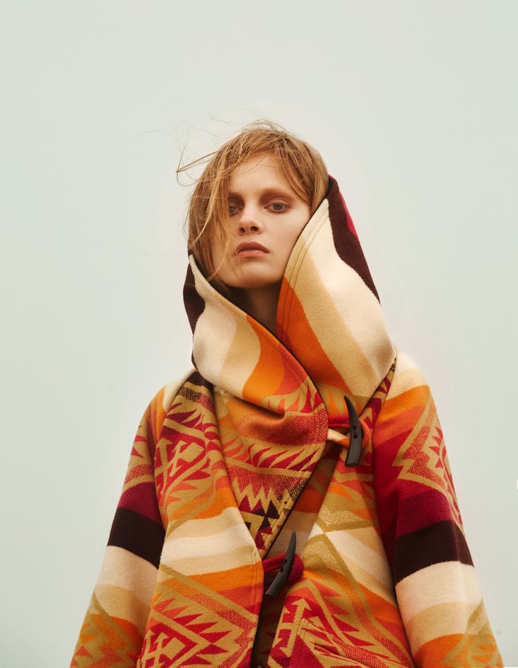 a young woman with long brown hair wears a colorful striped and patterned coat with a hood, posing for a portrait in a studio. she looks at the camera with a neutral expression. the coat is folded at the front and secured with a brown toggle. the background is a simple light grey.