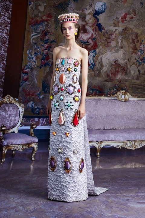 a woman stands in a room with intricate interior design elements. she is wearing a long silver gown with a jeweled top and a crown on her head. the dress is covered in jewels and the woman has a beautiful and glamorous look.  there is a tapestry on the wall behind her and a large ornate chair and sofa in the room.  the woman is a model and the setting is a studio for a fashion shoot.  the image is highfashion and  showcases the beautiful design and style of the woman's clothing.