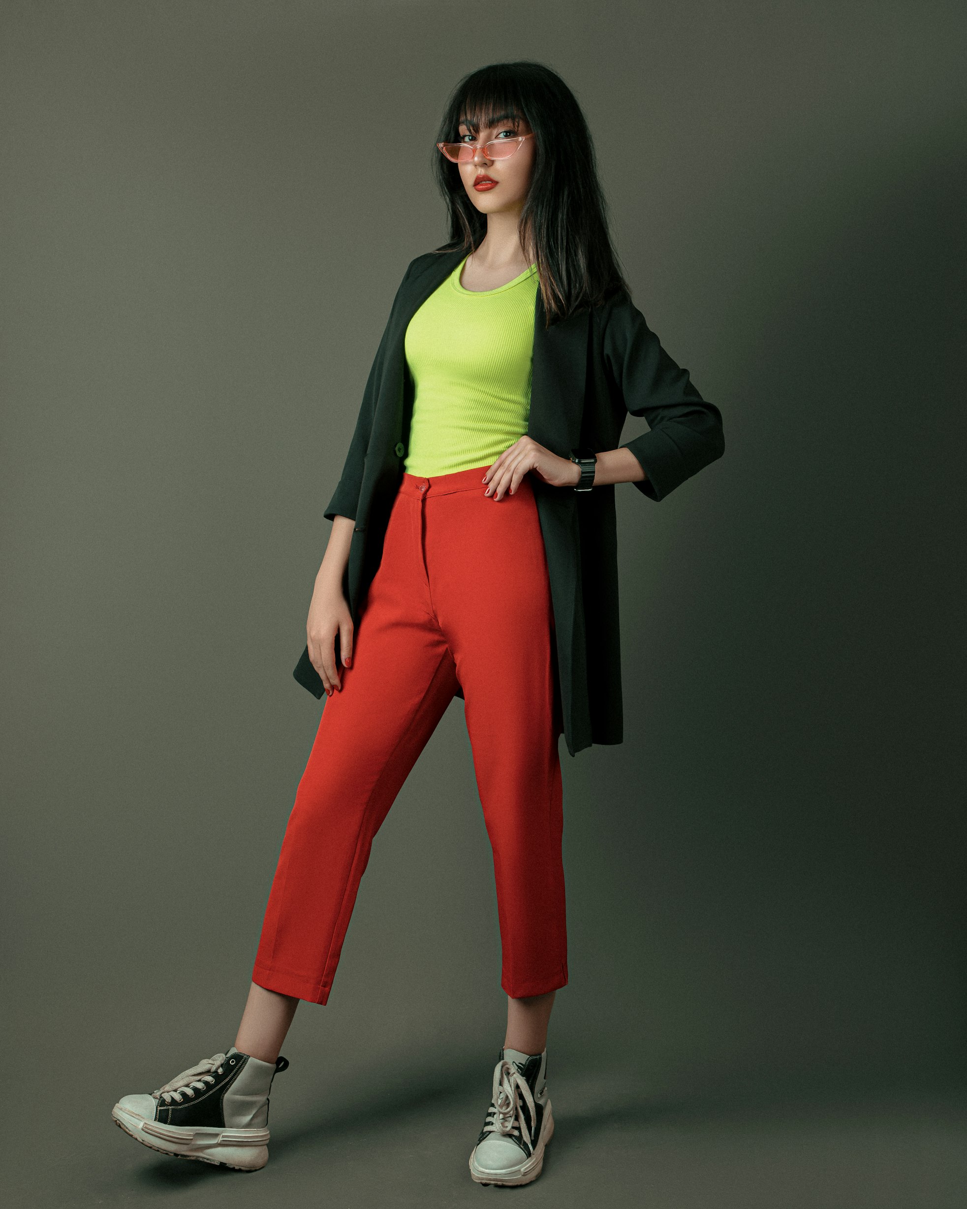a fulllength studio shot of a woman posing in a fashionable outfit. she is wearing red pants, a neon green shirt, a black jacket, and black and white sneakers. she is also wearing pink sunglasses and a wristwatch. the background is a solid gray color.