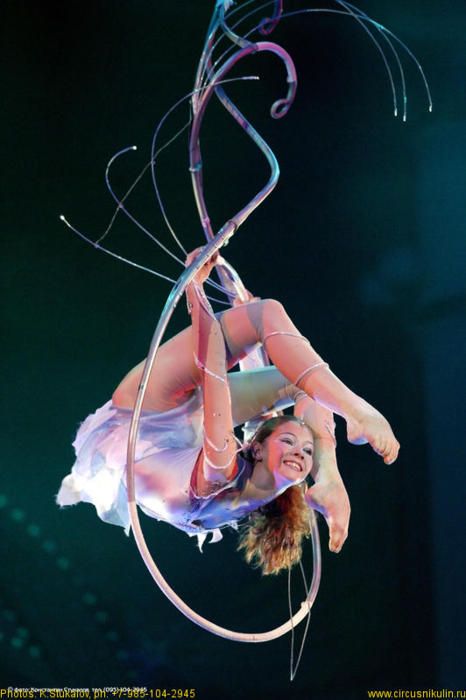 a woman is performing an aerial hoop routine. she is hanging upside down in the hoop, showing impressive flexibility and strength.  she is surrounded by  a steel hoop with glowing lights and intricate metal wires.  she is wearing a white and blue costume.  her long hair is flowing and she is smiling, enjoying her performance.