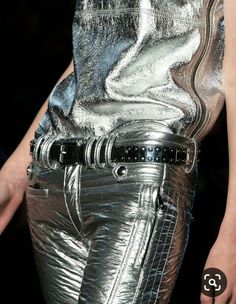 a woman in a silver, leather outfit poses for a photoshoot. the outfit includes silver pants, a silver top, and a silver leather belt. she is wearing a stylish, glamorous outfit that emphasizes her figure.