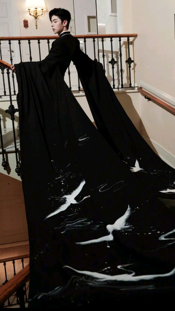 a man in a black suit with a long train decorated with white crane designs stands on a staircase. the dress extends from his arms and drapes down the stairs. a railing borders the staircase and a lamp is visible on the wall.