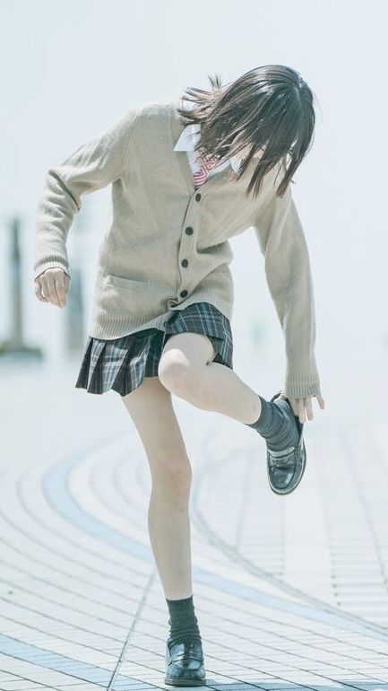 a young japanese school girl in uniform poses for a photograph. she is wearing a white shirt and a gray sweater over a plaid skirt, and black shoes and socks. she is standing in a street, looking down at