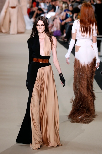 two fashion models are walking down the runway, showing off haute couture designs.  the model on the left is wearing a dress with a black and beige color scheme,  a brown belt, and black gloves.  the model on the right is wearing a white dress with brown feathers on the bottom of the skirt.  the background is blurred and features a large audience.