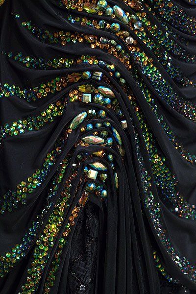 a closeup shot of a black dress featuring an array of colorful rhinestones and sequins. the fabric is draped in a way that creates pleats and gathers, showcasing the texture and embellishment. the rhinestones and sequins are arranged in patterns that add an elegant and luxurious feel to the garment.