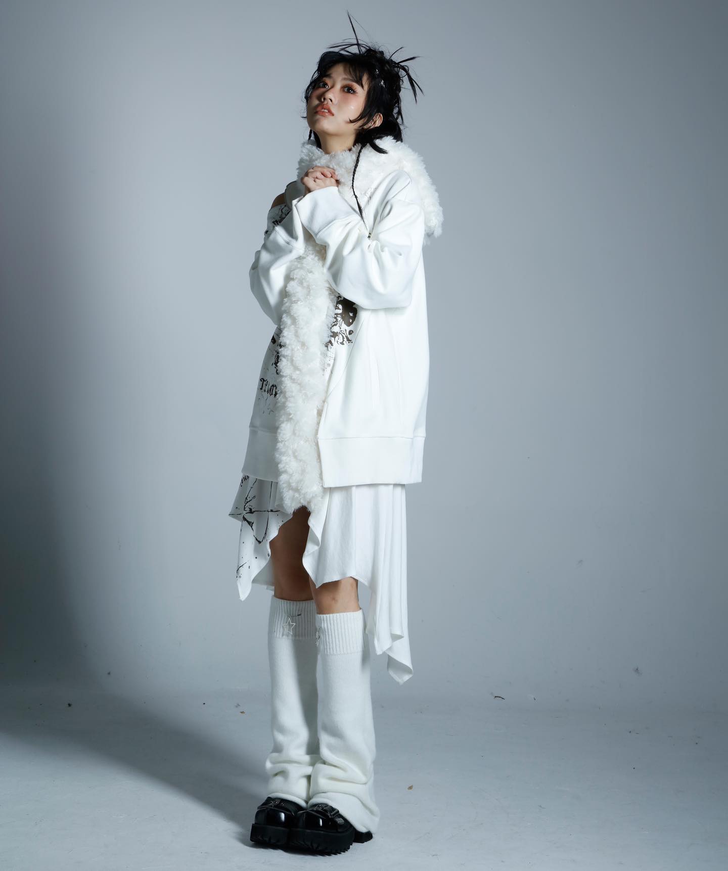 the image shows a young woman, possibly a model, posing in a studio against a light gray backdrop. she's dressed in an allwhite outfit composed of an oversized hoodie with a fluffy white fur trim, an asymmetrical skirt with graphic details, and tall white socks paired with black boots. her hair is styled in a slightly messy updo, adding to the overall grunge or alternative fashion aesthetic. the pose and styling suggest a photoshoot for a clothing brand or fashion publication.
