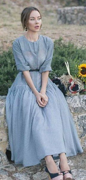a woman in a pretty blue gingham dress sits on a stone wall outdoors.  she is surrounded by sunflowers and greenery. the dress has a gathered skirt and elbowlength sleeves.  the overall mood of the image is peaceful and serene. she looks calm and relaxed.  the setting suggests a rural or countryside location.