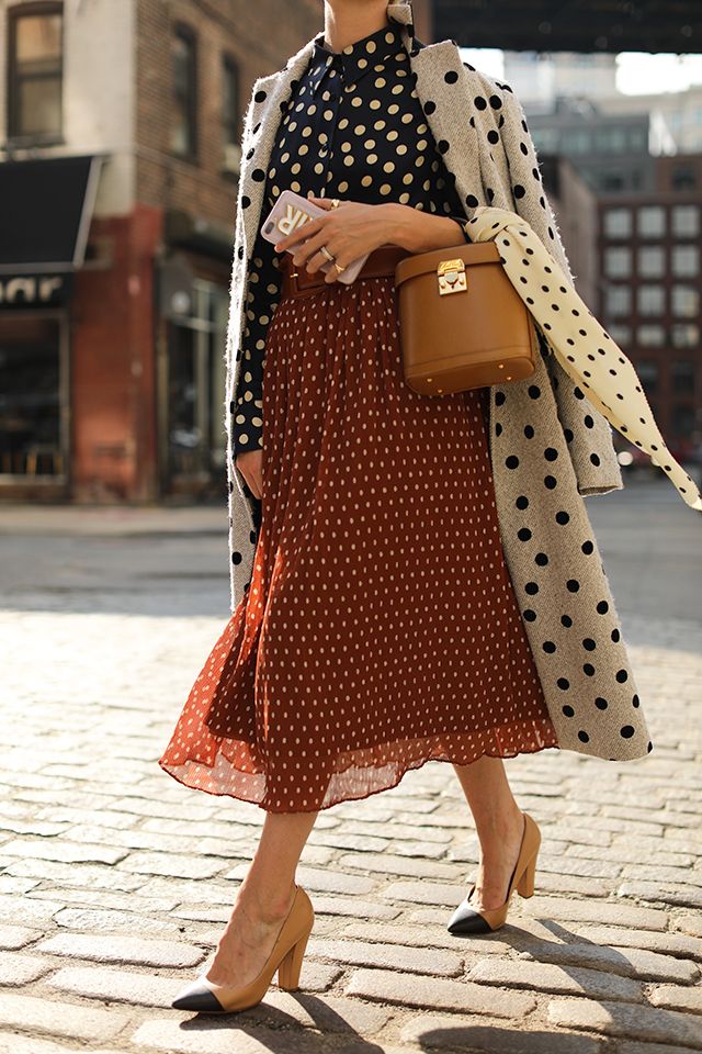 a stylish woman strolls down a city street, her outfit exuding autumnal charm. she is wearing a rustcolored polka dot midi skirt, a navy polka dot blouse, and a stylish gray polka dot coat.  the midi skirt is pleated, giving it a subtle elegance. her accessories include a tan structured handbag, and twotone shoes with a beige upper and black toe. the overall look is effortlessly chic, reflecting a current fashion trend. the backdrop of a brickpaved street and urban buildings enhances the image's sense of style and atmosphere.