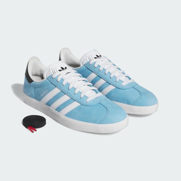 a pair of light blue adidas gazelle shoes with white stripes, black accents and white laces are displayed on a white background.  they appear to be new and unused.  a red shoelace is in the foreground.
