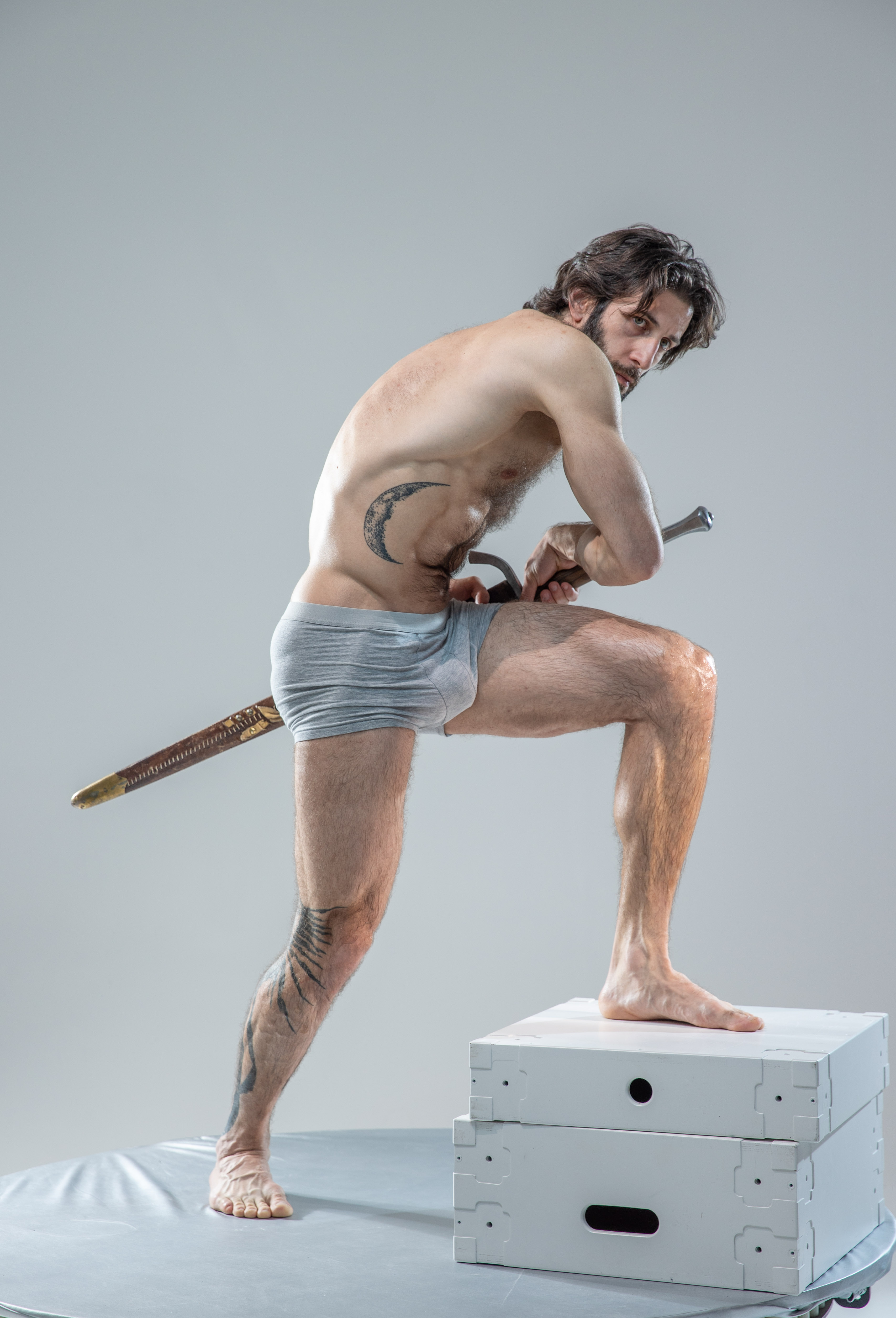 a man with long hair and a beard stands in a warrior pose on top of white boxes against a grey background. he is shirtless, wearing grey underwear and holding a sword with a gold and black hilt. he has a tattoo of a crescent moon on his side and a tattoo on his left leg. he is illuminated with soft studio lighting.